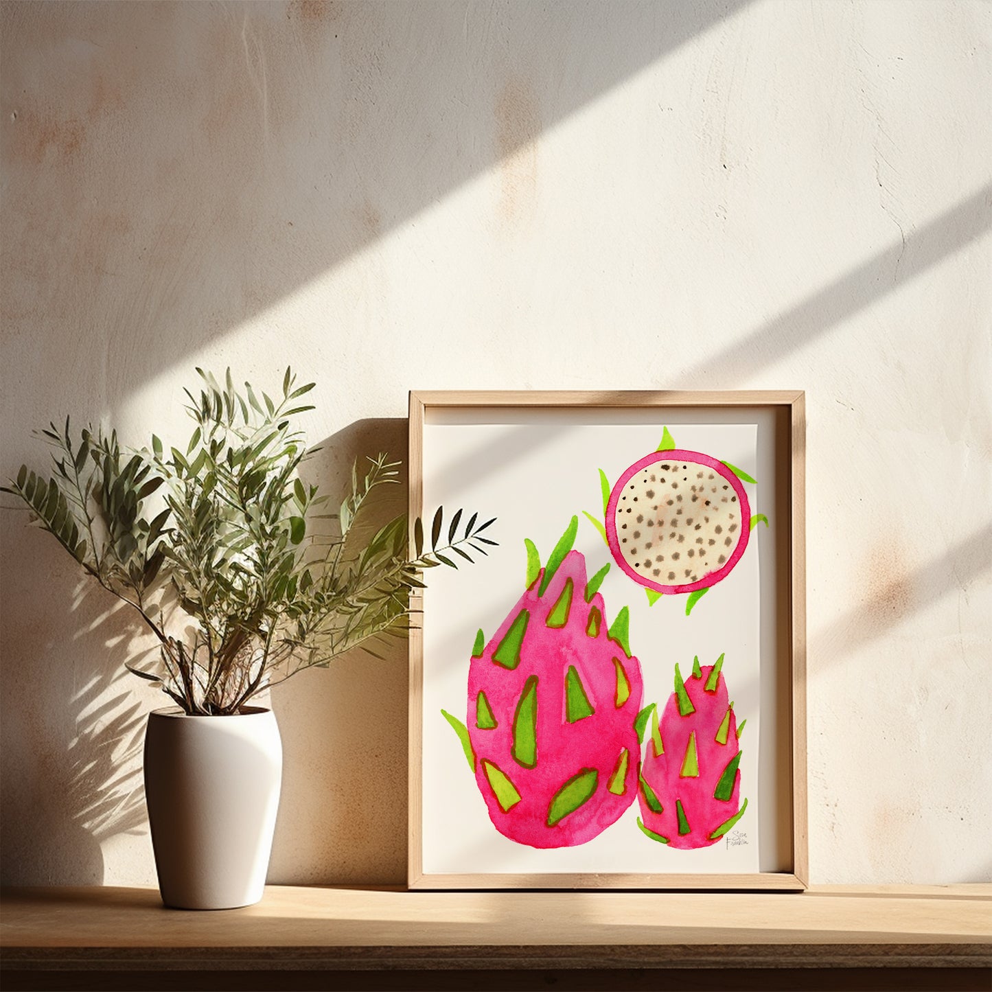 a framed watercolor painting of three dragon fruits, with a white vase of green leaves placed next to it on a wooden shelf.