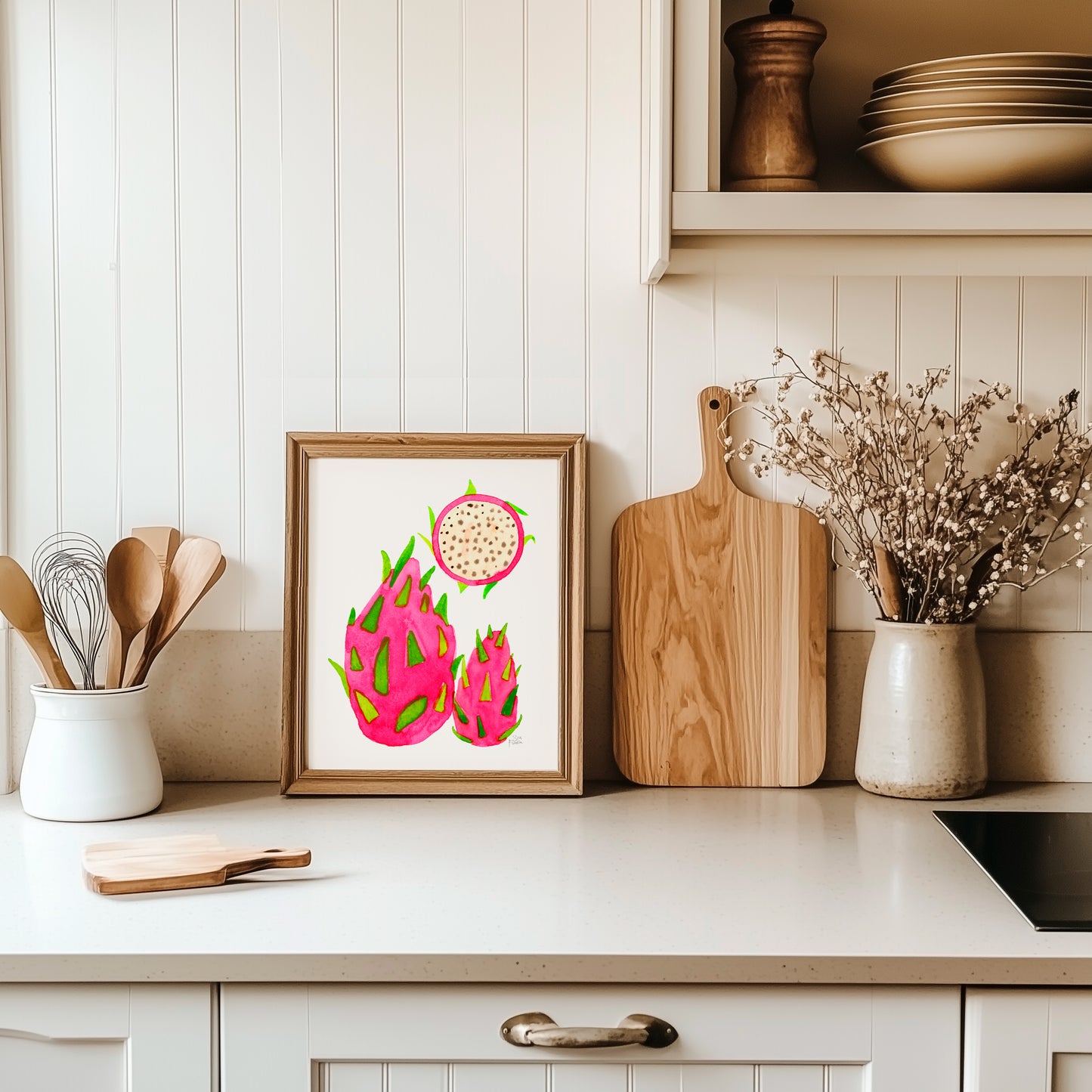a kitchen counter with a framed print of three dragon fruits, wooden cutting boards, and a vase of flowers.