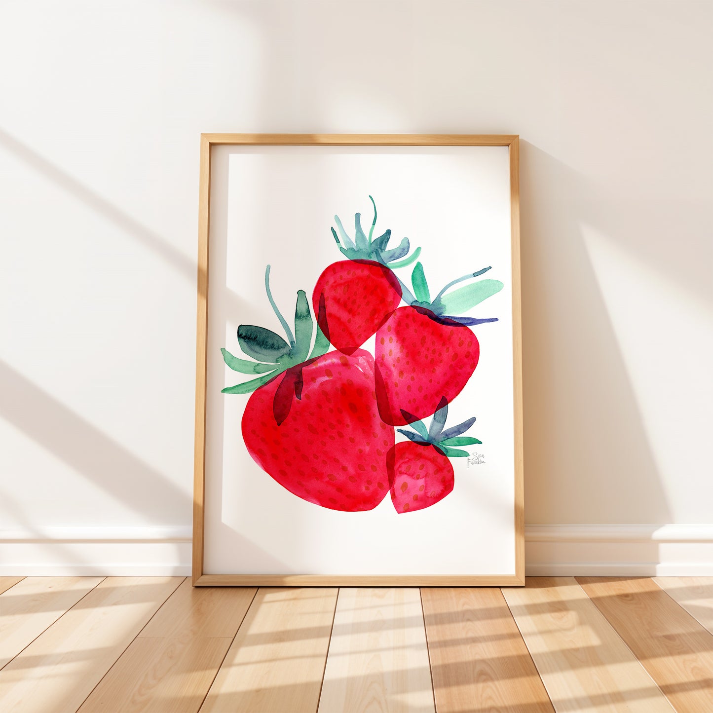 A framed watercolor painting of red strawberries is displayed on a white wall in a room with a wooden floor.