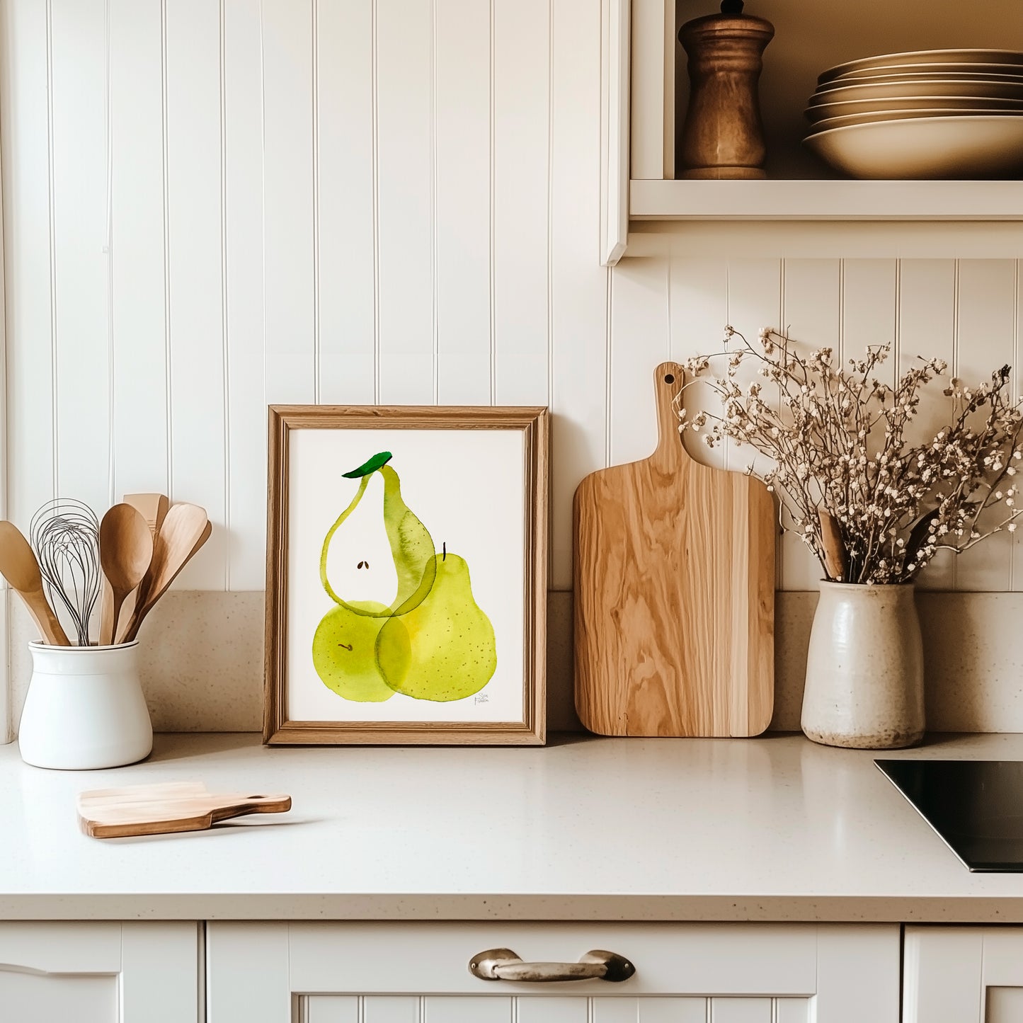 a kitchen counter with a framed picture of two green pears, a wooden cutting board, and a vase of flowers.