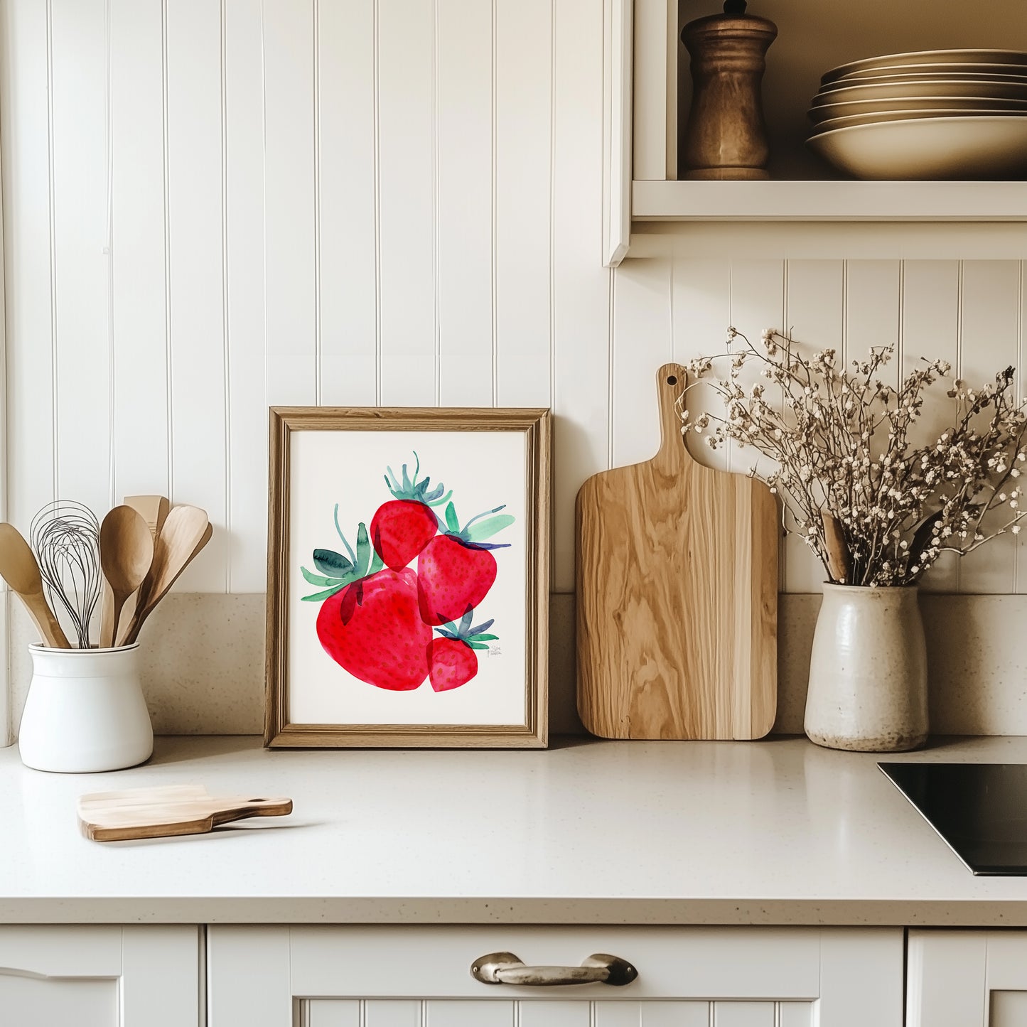 a kitchen counter with a framed picture of red strawberries, wooden cutting boards, and a vase of white flowers.