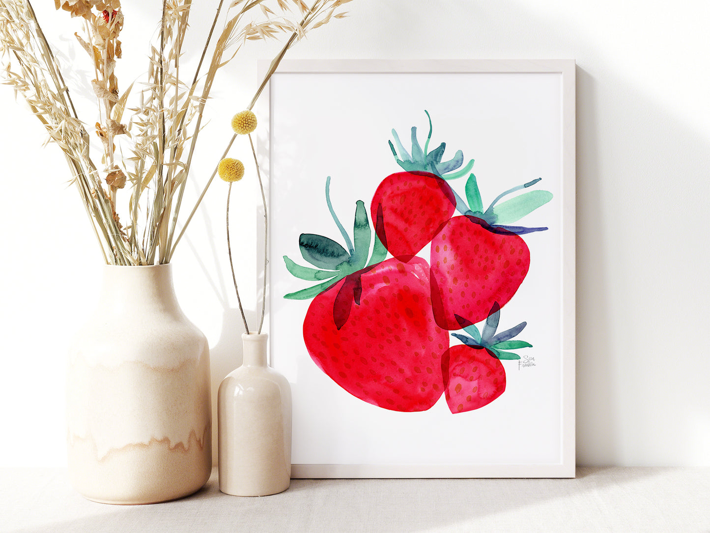 a white vase with dried flowers and a framed painting of red strawberries on a white wall.