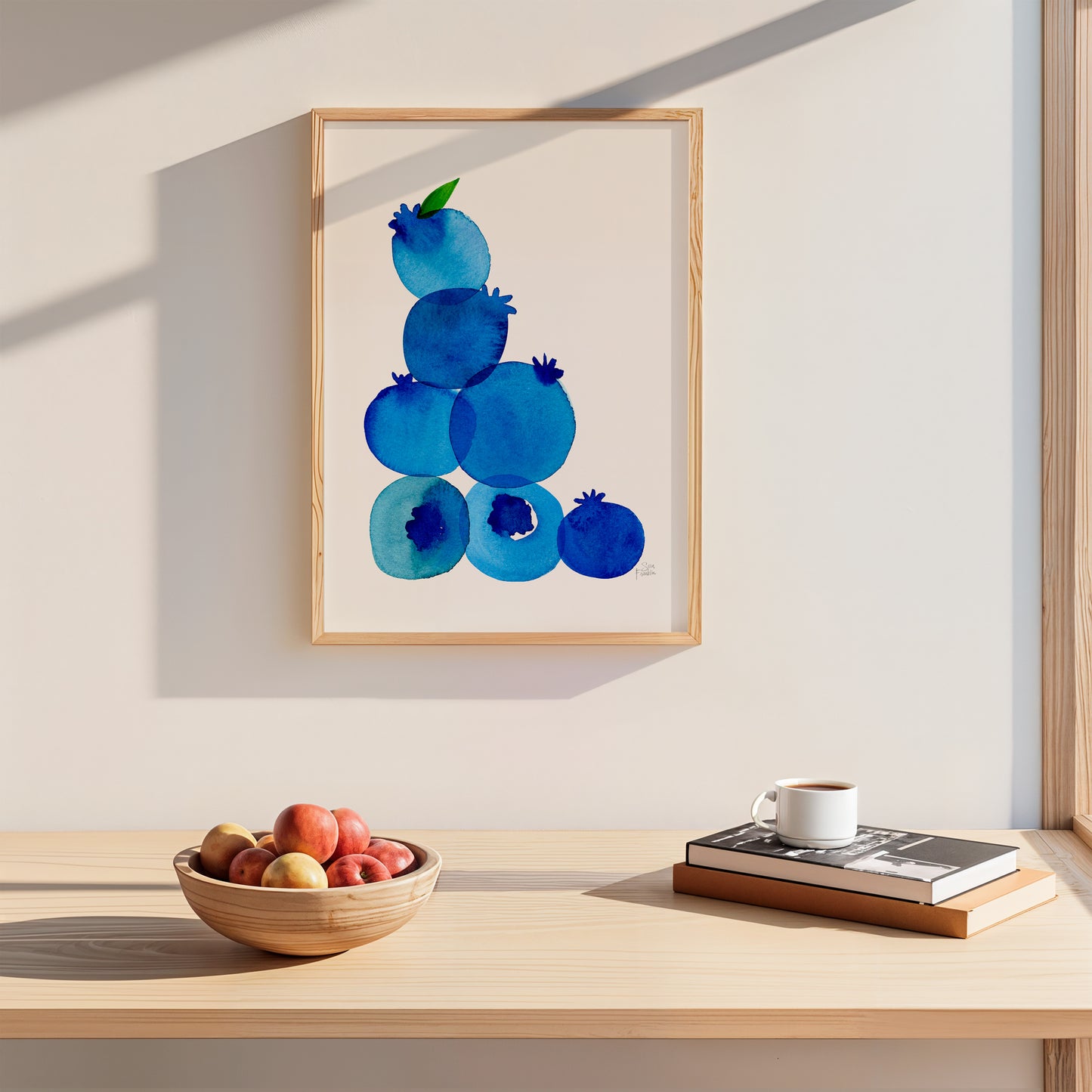 a wooden table with a bowl of fruit, a cup of coffee, and a stack of books. Above the table, there is a framed painting of blueberries.