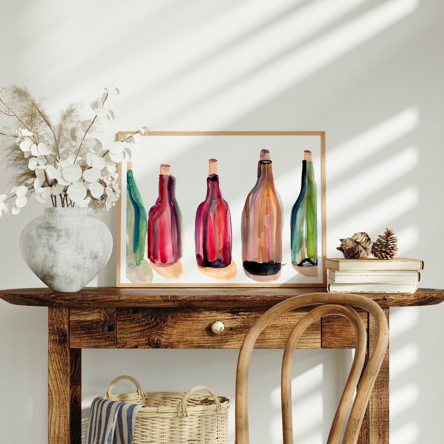 a wooden table with a framed painting of colorful bottles on it, a white vase with white flowers, a stack of books, and a woven basket.