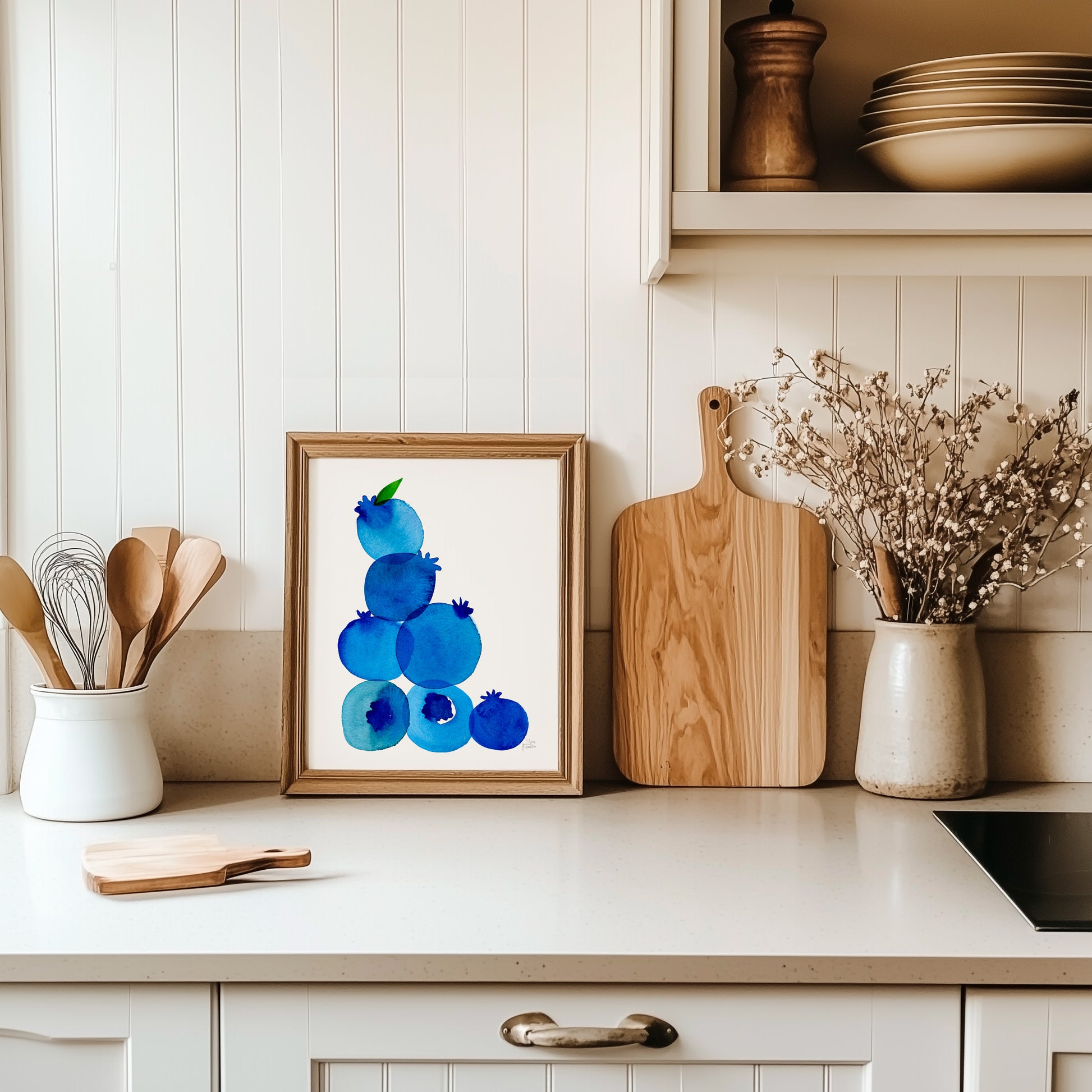 a kitchen counter with a framed artwork of blueberries, wooden cutting boards, and a vase of flowers.