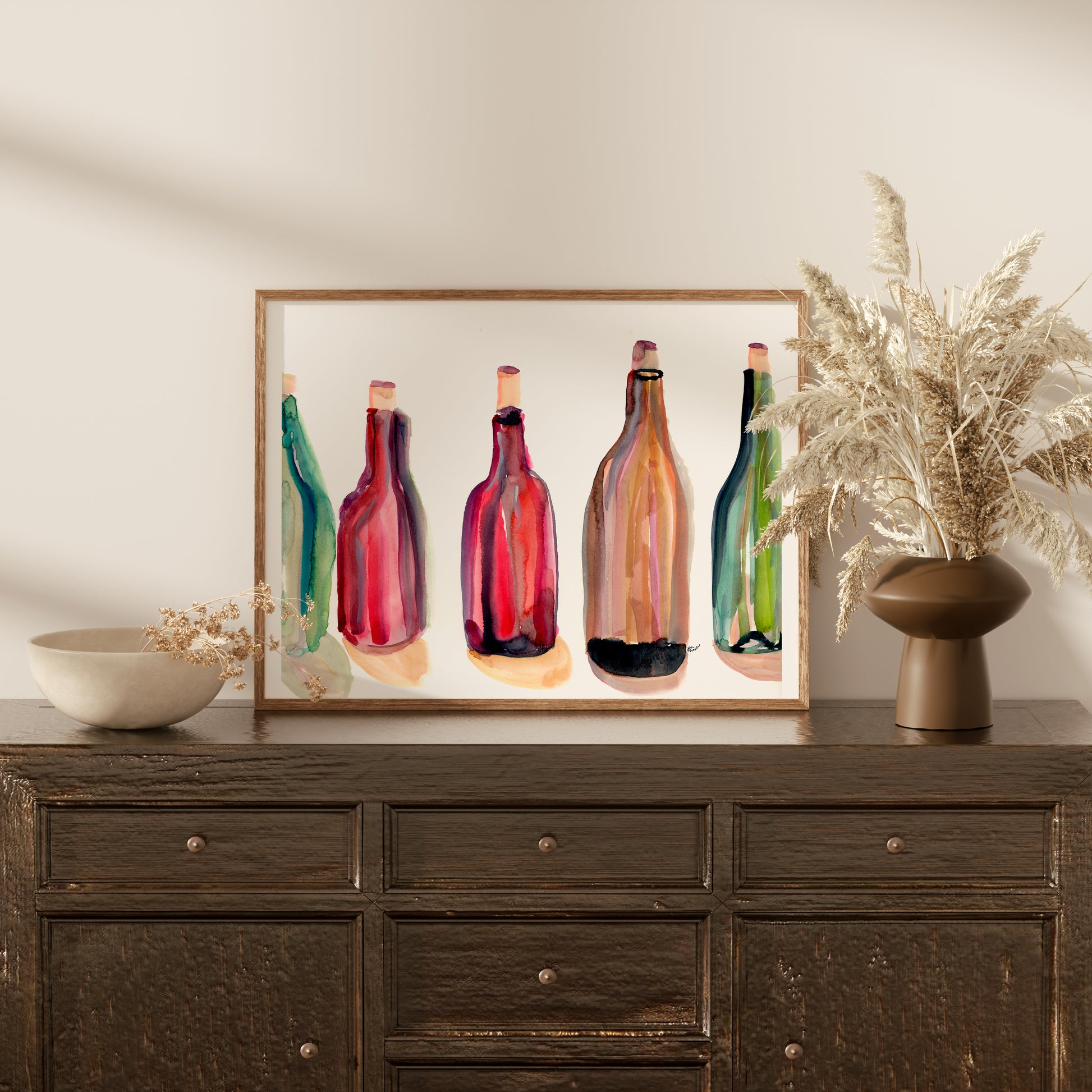 a wooden dresser with a framed painting of colorful wine bottles on top. The dresser has a vase of dried flowers and a bowl on it.